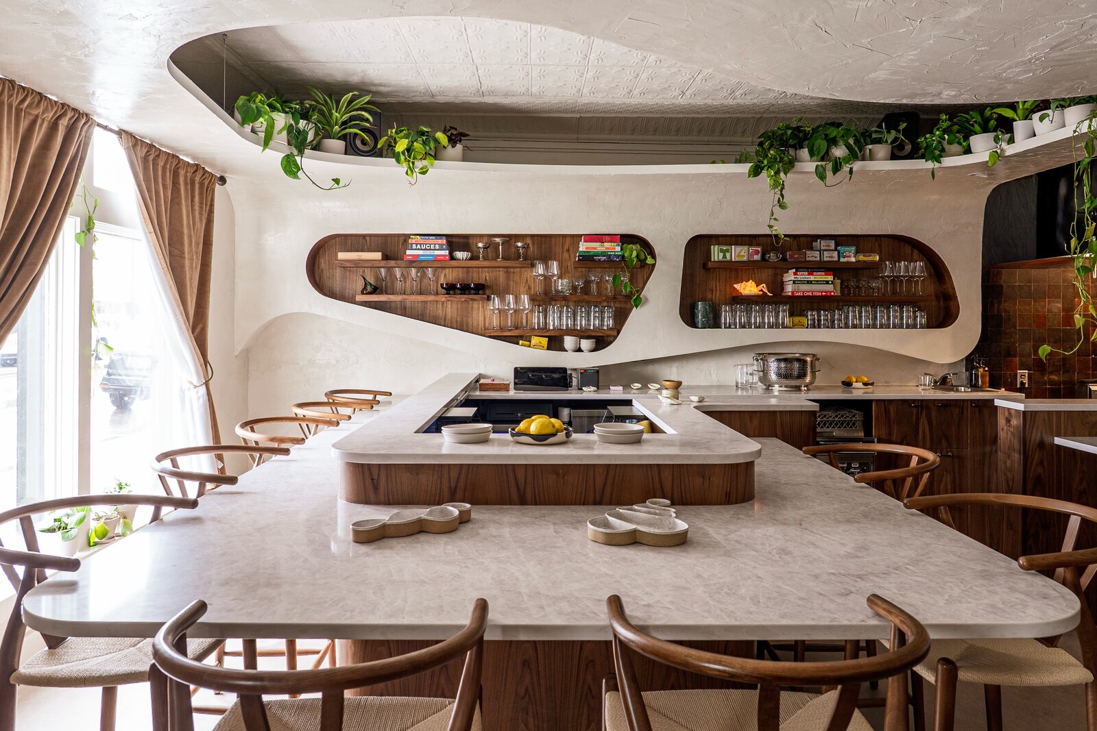 Umbo's dining room — a curved plaster ceiling with trailing plants, wavy walnut shelving of bottles and glassware, and a marble bar lined with wishbone chairs
